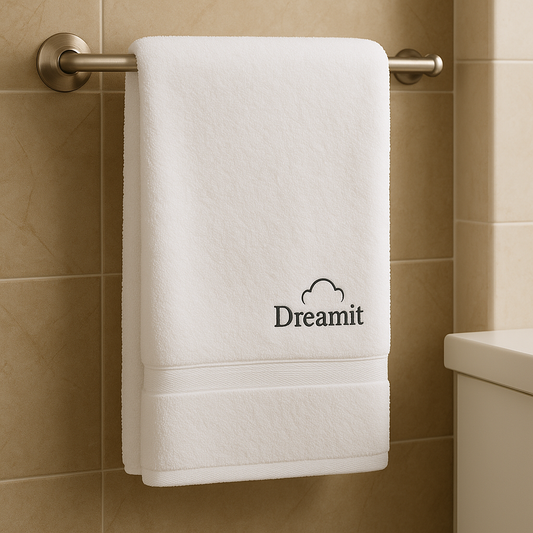 מגבת גוף לבנה עבה ואיכותית תלויה על מתלה מתכתי בחדר אמבטיה מוסדי, עם רקע של אריחים בהירים. לוגו Dreamit המקורי מופיע בגוון אפור כהה עם סמל ענן פתוח, ממוקם בפינה הימנית התחתונה של המגבת – מראה מקצועי, נקי ומתאים לבתי מלון, קליניקות ומוסדות בריאות.