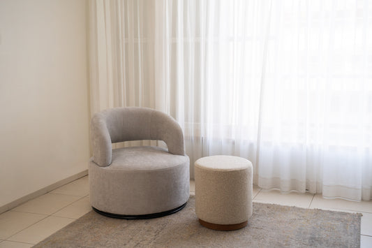 כורסה אפורה עם הדום שמנת בפינת סלון מודרנית ליד וילונות לבנים.