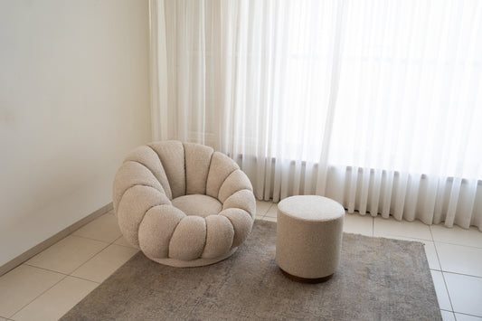 כורסה מעוצבת מבד בוקלה בצבע בז׳ עם הדום תואם ליד חלון ווילון לבן – פינת ישיבה מודרנית.